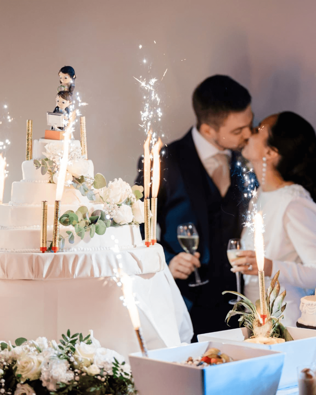 Couple s'embrassant devant un gâteau de mariage blanc décoré de fleurs et de cierges magiques.