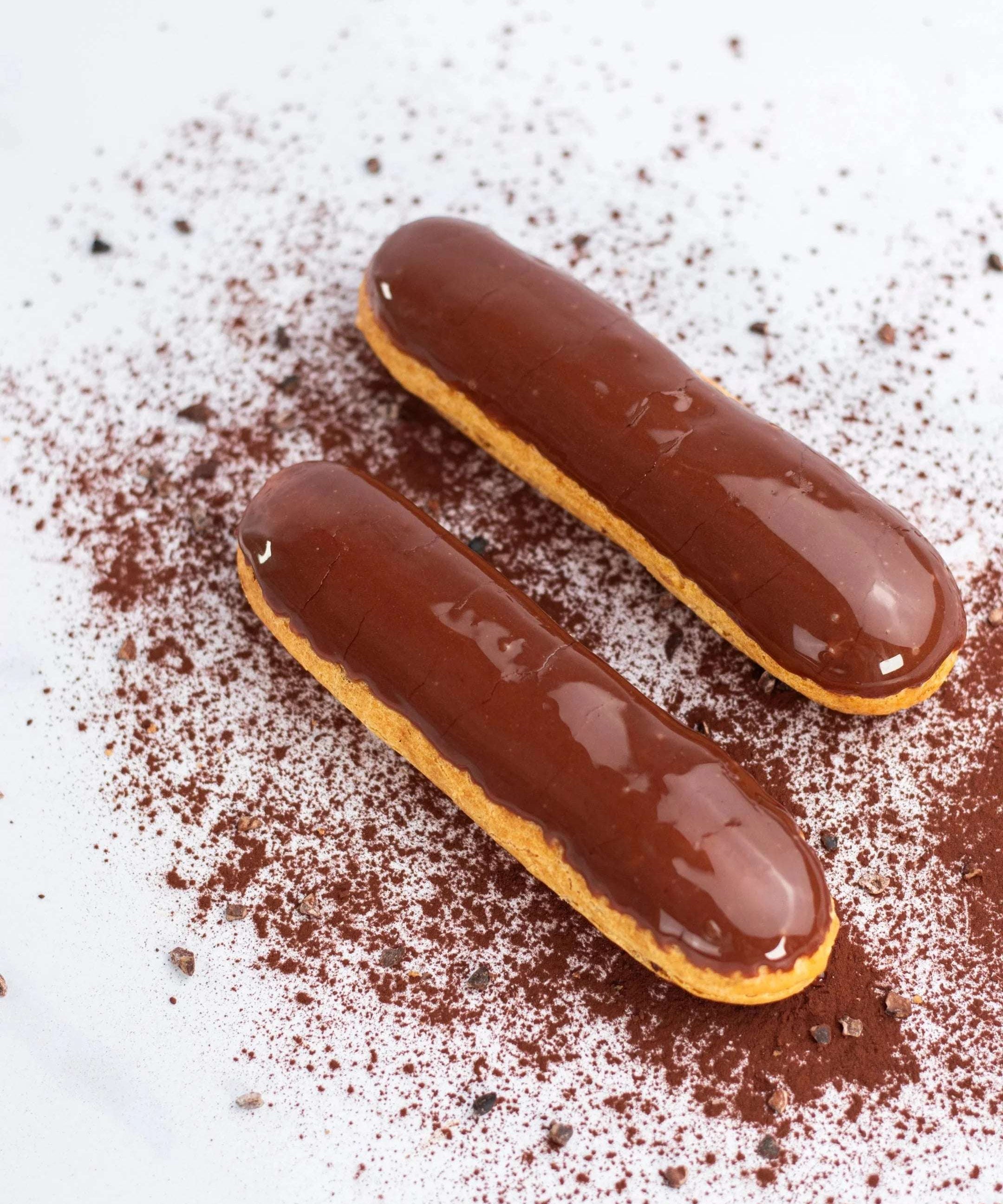 Deux éclairs au chocolat Maison Vatelier sur un fond blanc saupoudré de cacao.
