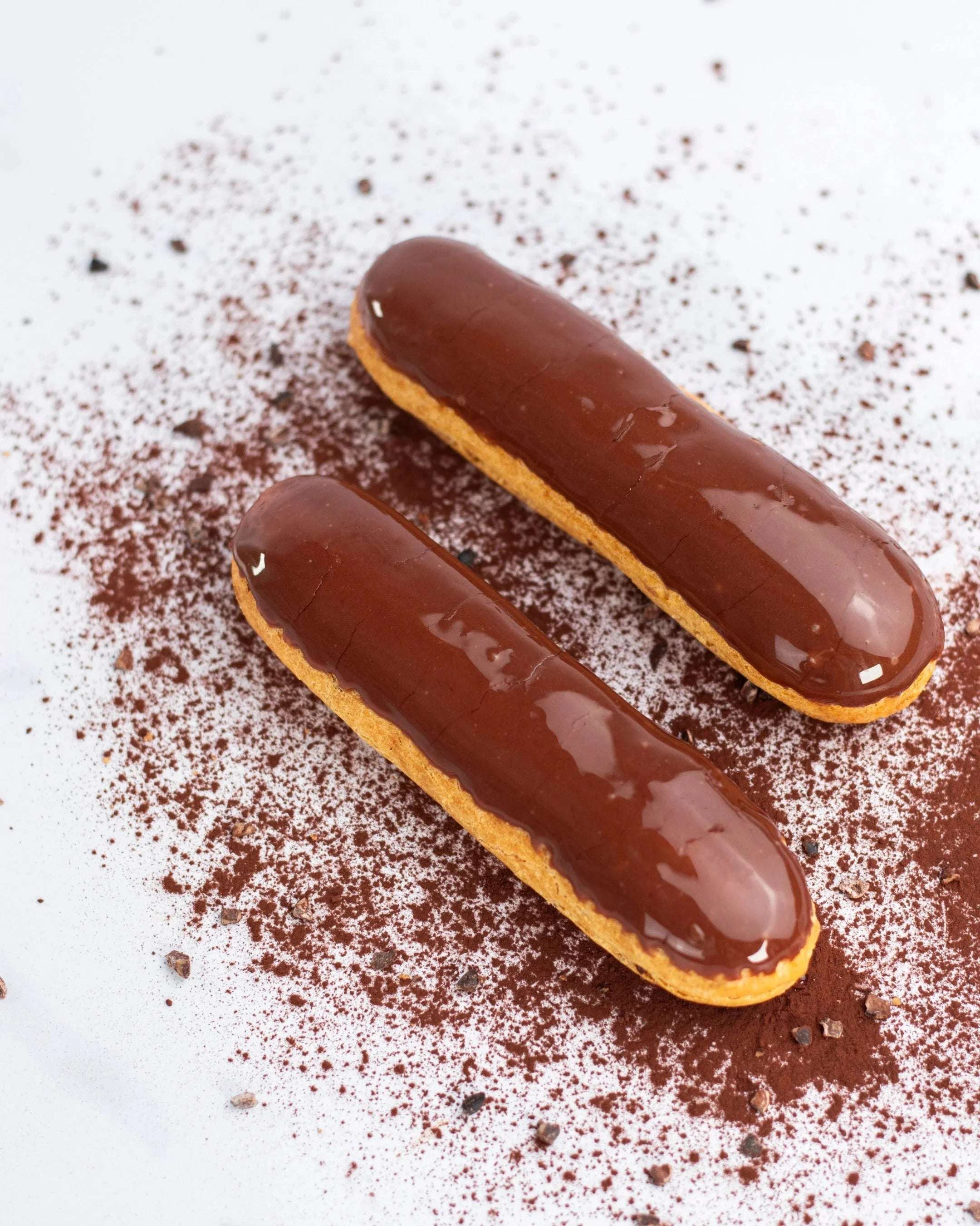 Deux éclairs au chocolat Maison Vatelier sur un fond blanc saupoudré de cacao.