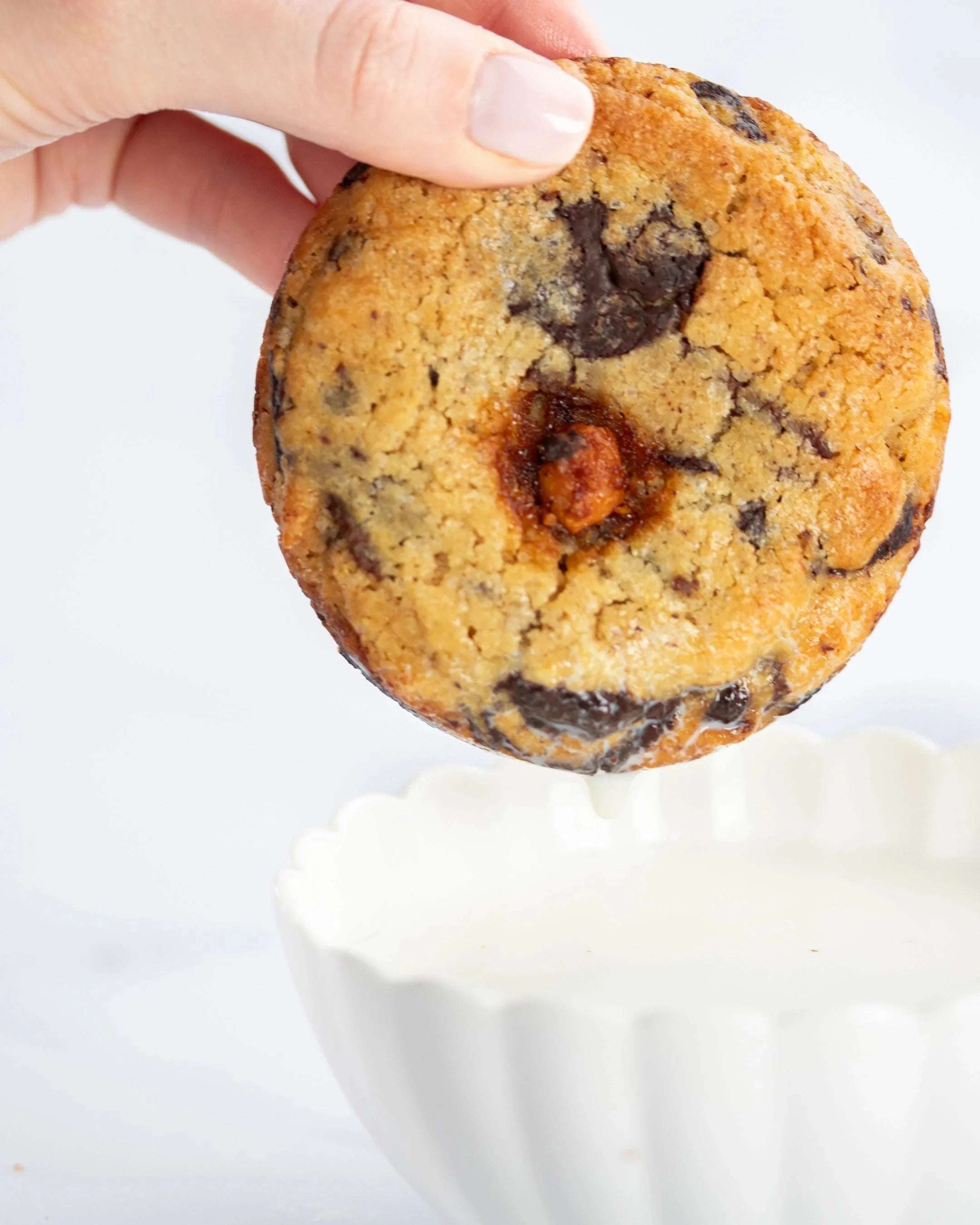Main tenant un cookie 3 chocolats Maison Vatelier au-dessus d'un bol de lait blanc.