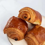 Trois pains au chocolat feuilletés et dorés Maison Vatelier sur un plateau blanc.