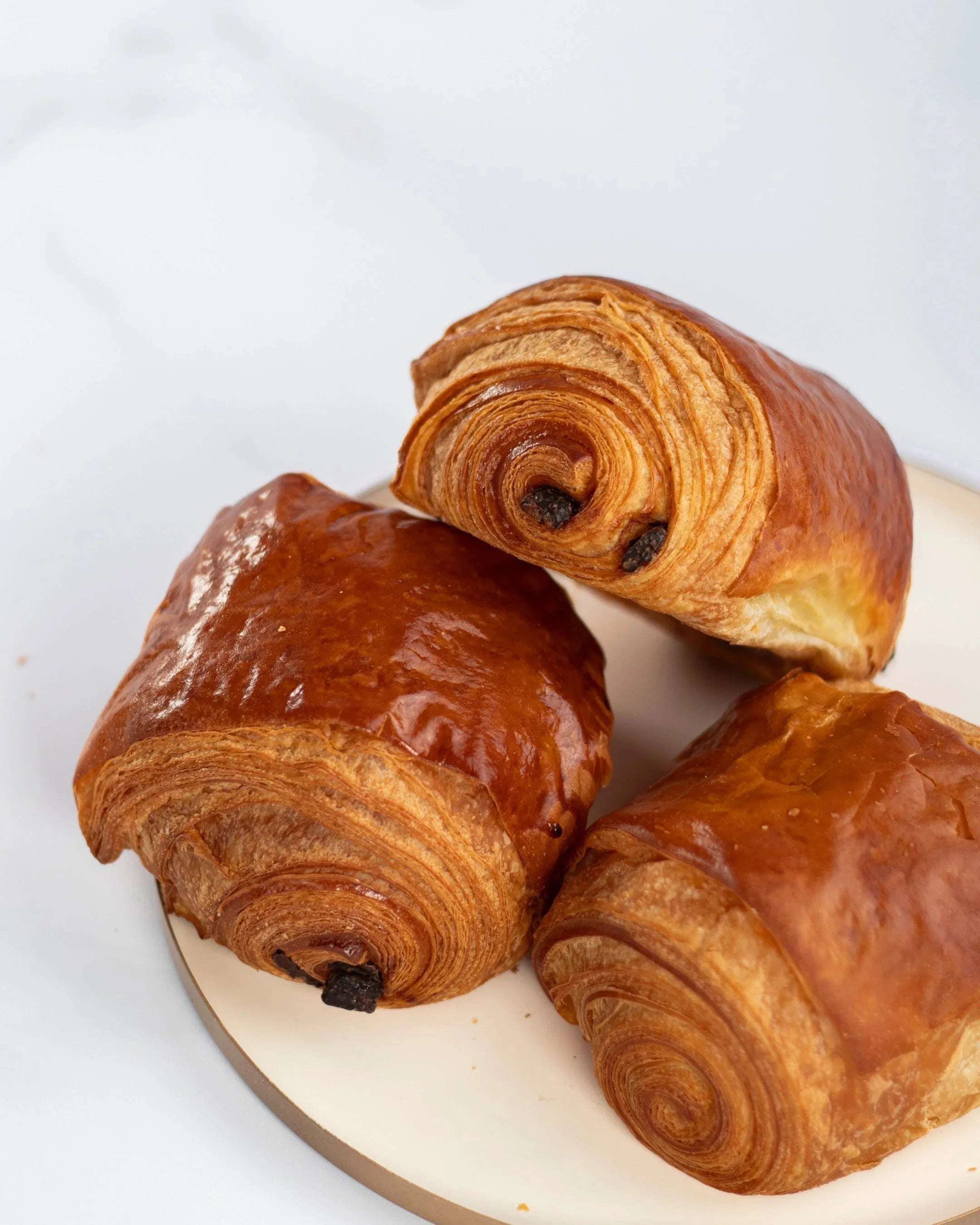 Trois pains au chocolat feuilletés et dorés Maison Vatelier sur un plateau blanc.
