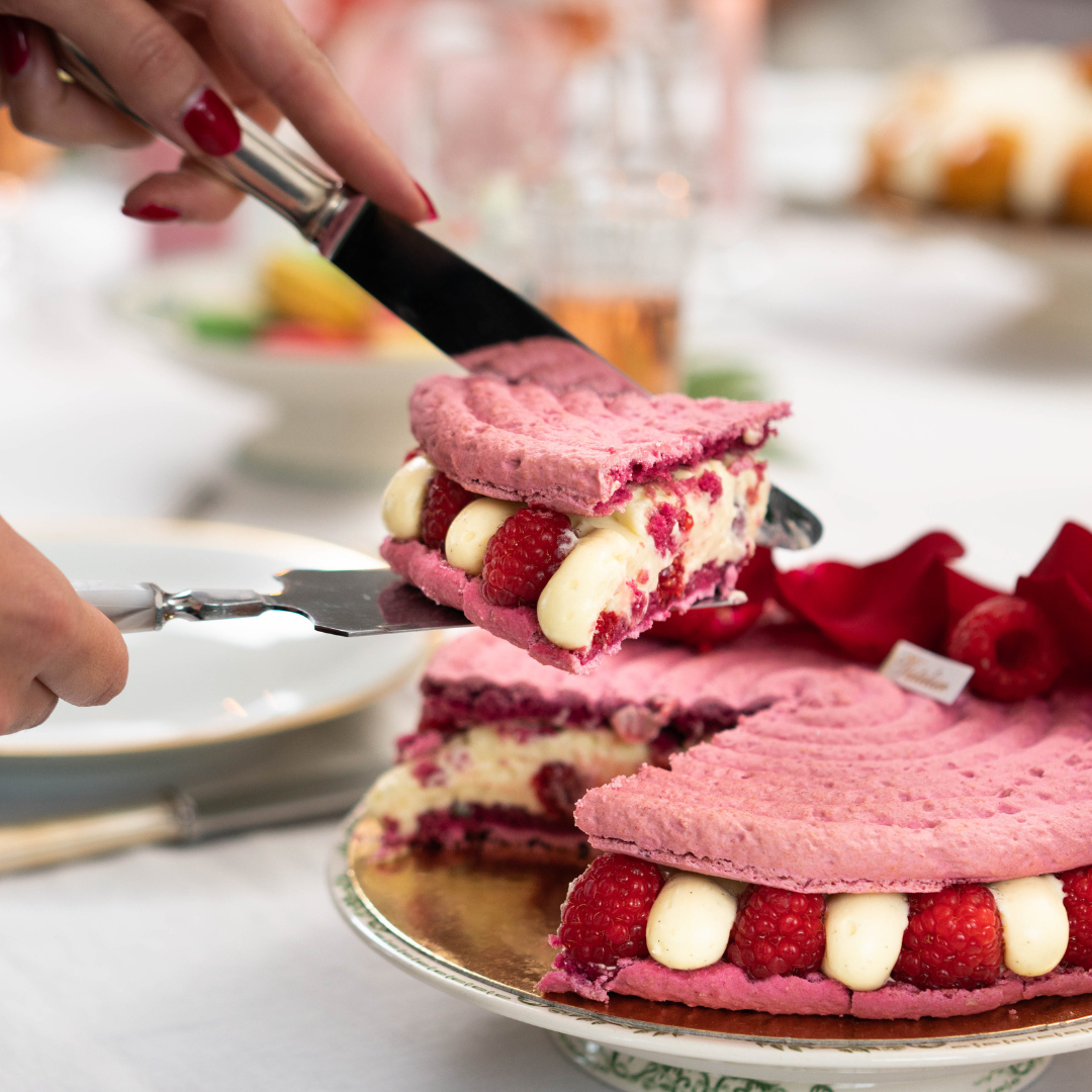 Une main servant une part de gâteau mousse framboise et crème sur un plateau blanc.