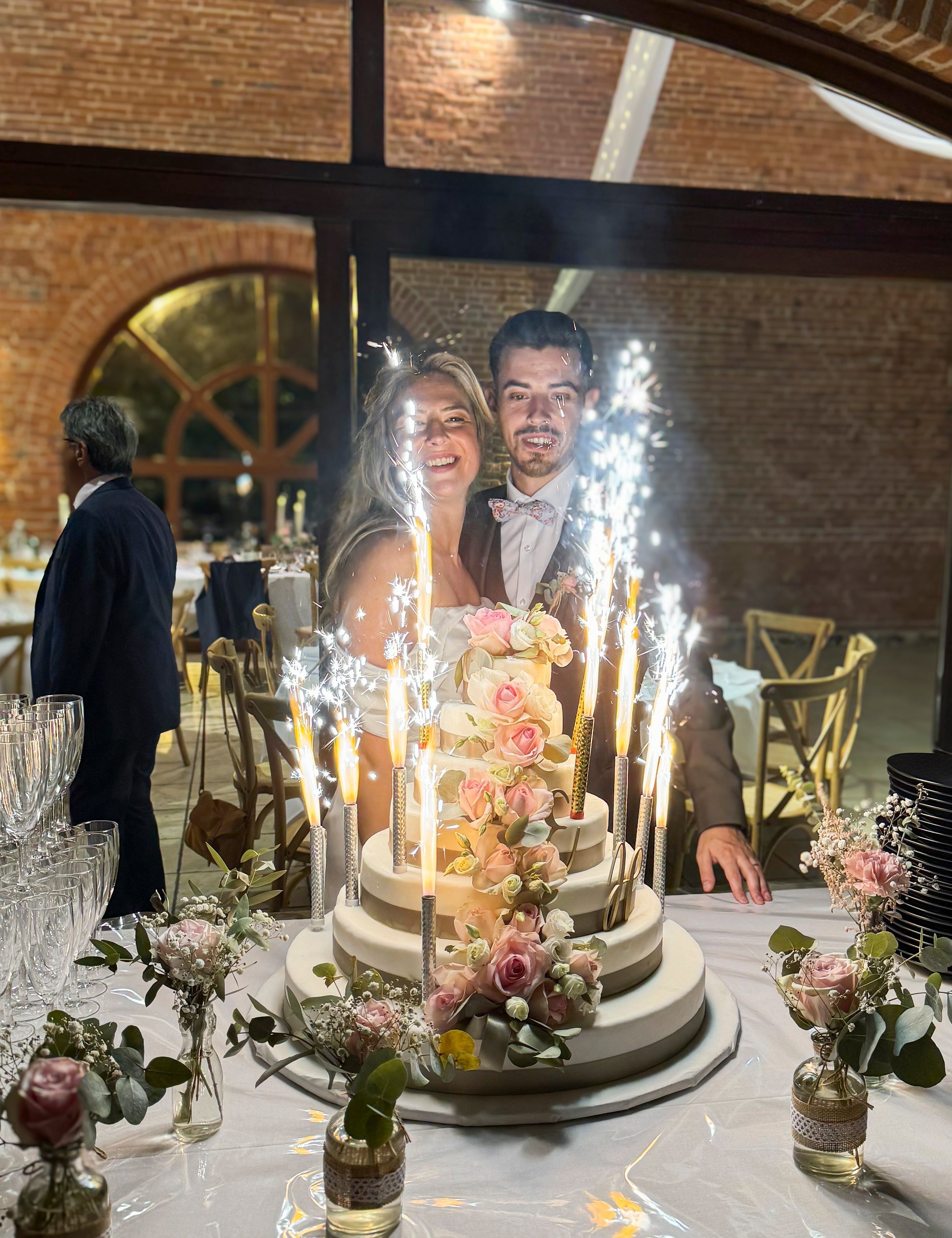 Couple souriant devant un gâteau de mariage à étages décoré de fleurs et de cierges magiques.