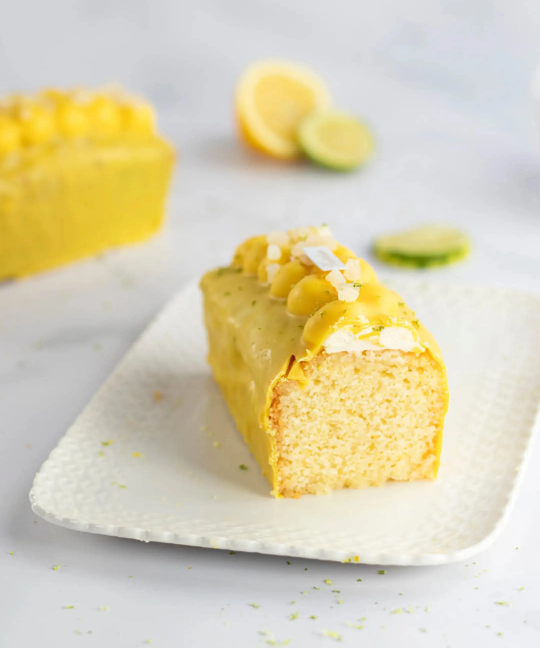 Tranche de cake au citron Maison Vatelier avec glaçage jaune et zestes sur assiette blanche.
