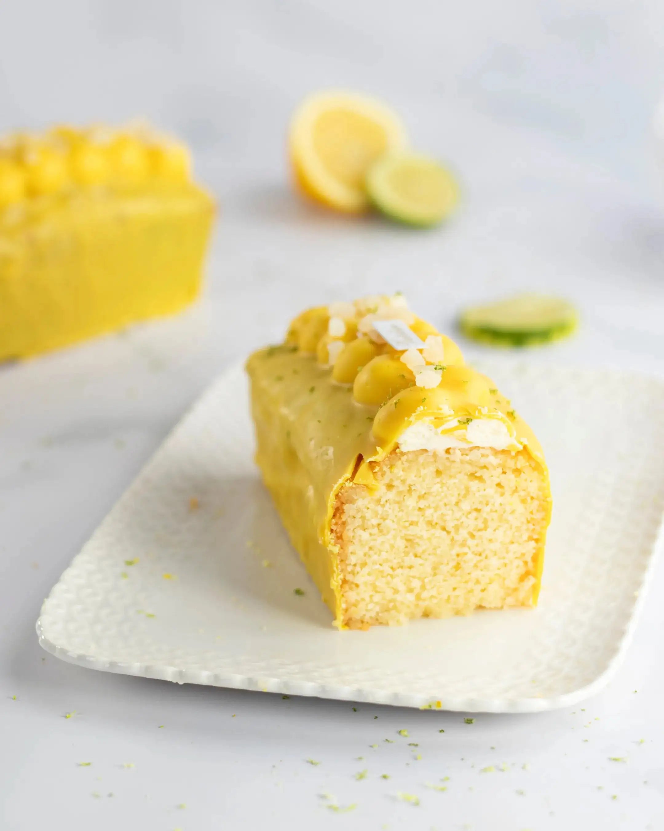 Tranche de cake au citron Maison Vatelier avec glaçage jaune et zestes sur assiette blanche.