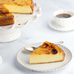 Part de flan vanille Maison Vatelier sur une assiette blanche avec le gâteau entier en arrière-plan.